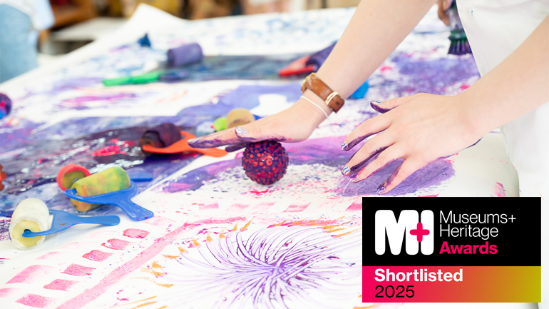 Two hands covered in paint. One hand is rolling a ball over a large sheet of white paper. Museum + Heritage Awards logo is featured in bottom right corner of the image.