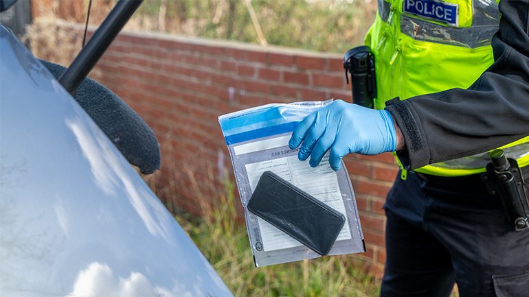 Police officer handles a mobile phone in an evidence bag beside a car