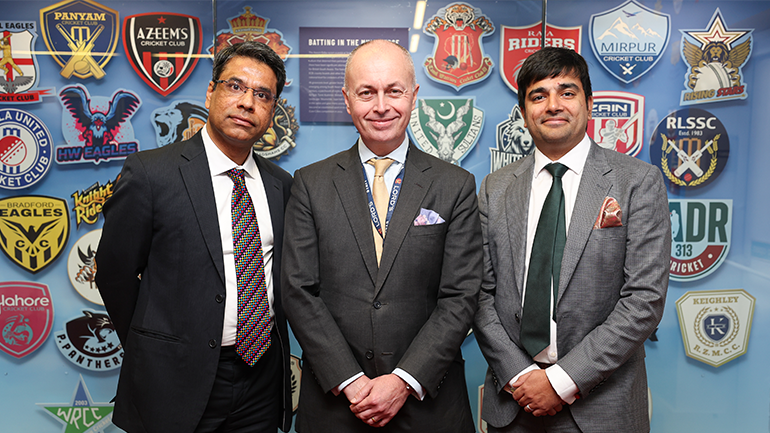 Three men in suits stand in front of a display wall covered with colourful cricket club crests and logos