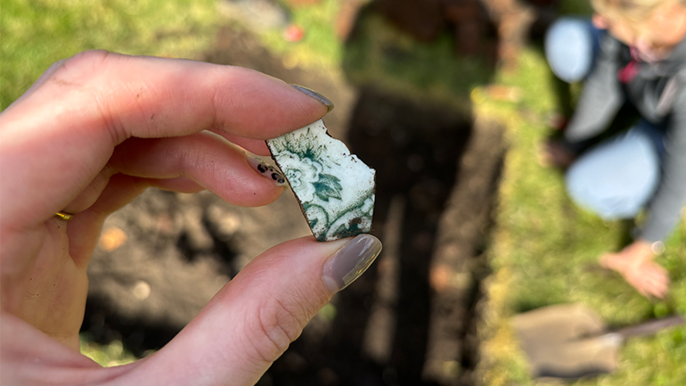 Close-up of a hand holding a small decorated pottery fragment, with an excavation trench and a crouching archaeologist blurred in the background