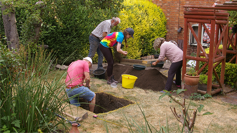 Four people take part in an archaeological dig in a back garden, with one person working inside a square test pit while others sift soil nearby