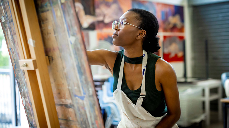 Person painting at an easel in an art studio, wearing an apron and glasses