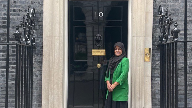 Person standing outside the front door of 10 Downing Street, wearing a green jacket and dark headscarf.