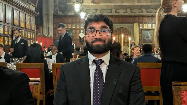 Person in a suit and tie seated at a formal dinner in an ornate hall.