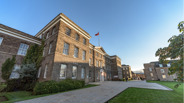 Exterior view of a large historic brick university building on a clear day. A paved pathway leads to the main entrance, with green lawns and landscaped planting on either side. A flag flies from the roofline, and additional campus buildings are visible in the background beneath a bright blue sky.