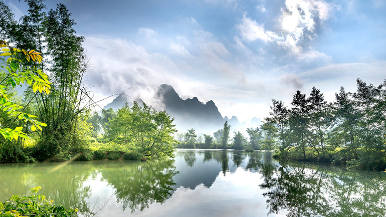 Peaceful river scene with mountain reflections and lush greenery under a bright sky. Sourced from Pexels.