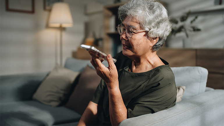 Older person seated on a sofa, speaking into a smartphone in a home setting.