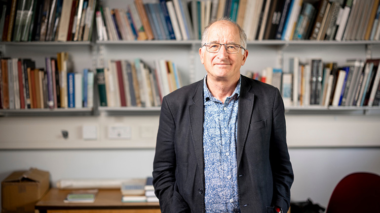 Person standing in front of bookshelves in an office or study, wearing glasses, a patterned shirt and a dark jacket.