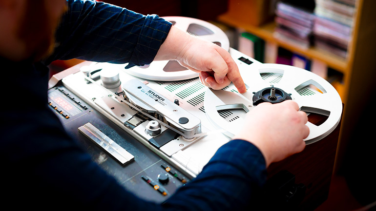 Person threading or adjusting tape on a reel-to-reel tape recorder, with large tape reels visible on the machine.