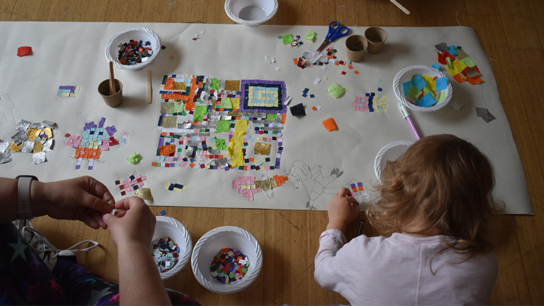 Children creating a colourful mosaic artwork from small paper pieces spread across a large sheet on the floor.
