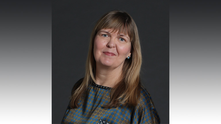 Studio portrait of Alice Donald with shoulder-length hair, wearing a patterned top, against a plain dark background.