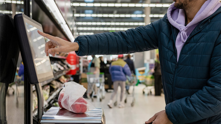 Person using a self-checkout machine in a supermarket, scanning items while a bag of produce sits on the scale.