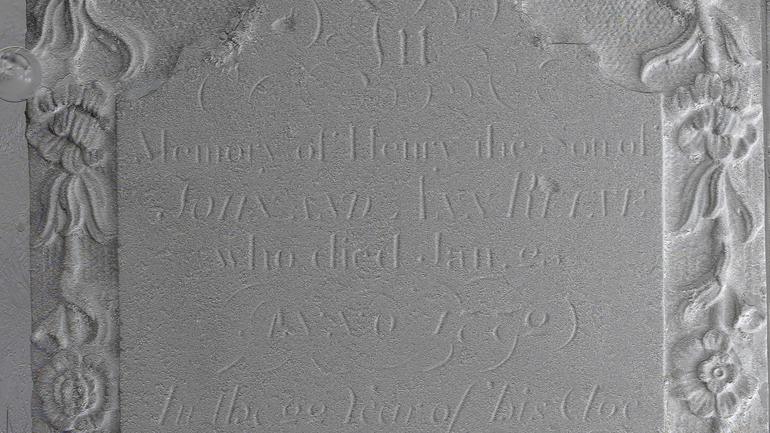 Close-up of an enhanced gravestone showing faint engraved text and decorative floral carvings along the sides.
