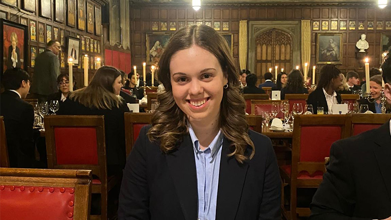 Person seated at a dining table in a wood‑panelled hall, with candles and other diners visible in the background.