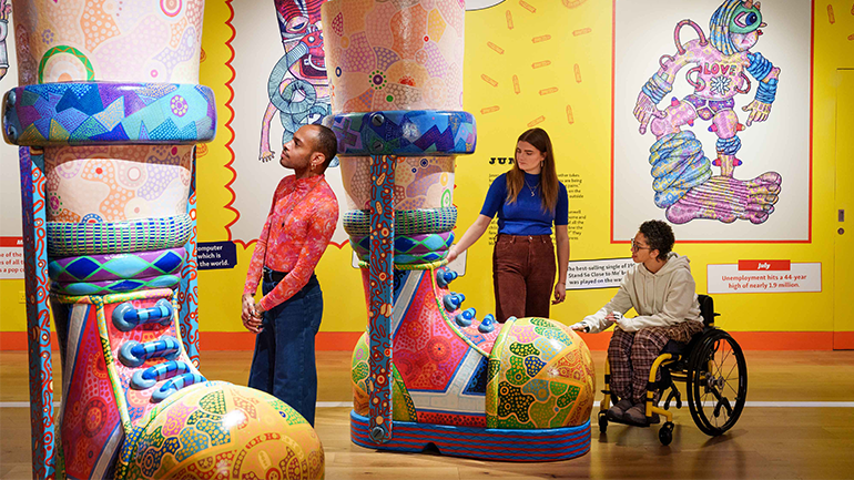 An indoor art exhibit with large, colourful patterned boot sculptures being explored by three people, including one using a wheelchair. A bright yellow illustrated wall with a character labeled “LOVE” forms the backdrop.