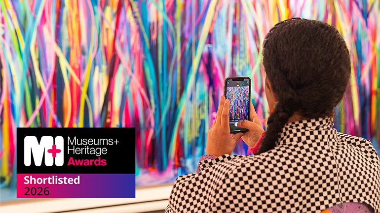 Person in a black-and-white checkered top photographs a brightly coloured abstract artwork in a gallery, with a “Museums + Heritage Awards – Shortlisted 2026” badge in the bottom left