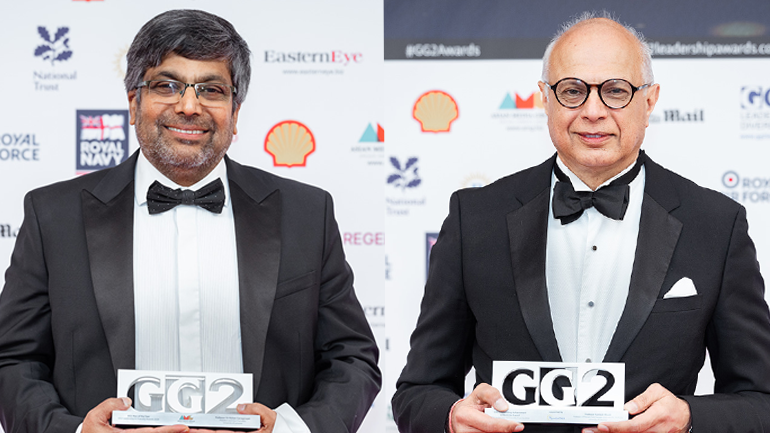 Two men in black tie stand in front of a branded awards backdrop, each holding a GG2 award and smiling at the camera.
