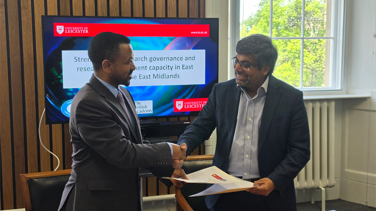 Two men in suits shake hands while one holds a document, standing in front of a University of Leicester presentation screen in a meeting room.