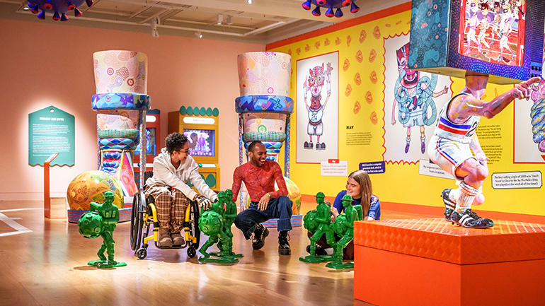 Three people chat on the floor of a colourful museum gallery beside small green sculptures, with large playful artworks and bright exhibition displays around them
