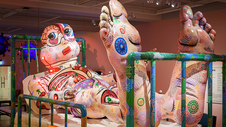 Large colourful sculpture of a reclining human figure with oversized feet and exposed anatomical details displayed in a museum gallery behind low barriers
