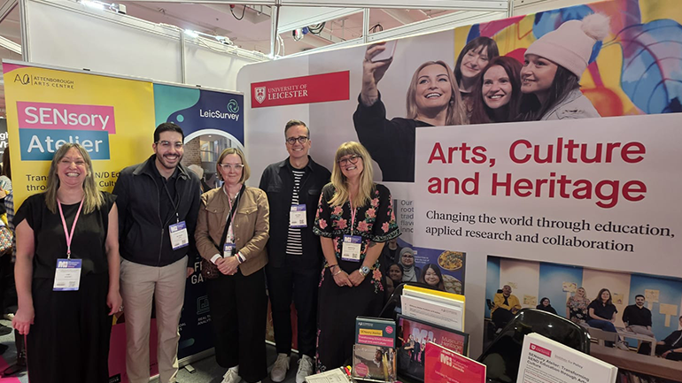 Five people stand smiling at a University of Leicester exhibition stand promoting arts, culture and heritage research, surrounded by banners, brochures and display materials.