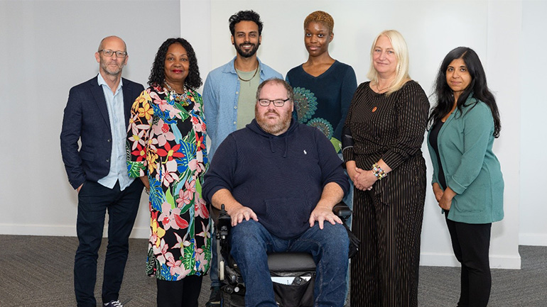 Group photograph of the Attenborough Arts Centre Advisory Board showing seven board members standing and seated together in a bright indoor space. One board member is seated in a powered wheelchair in the centre, with six other board members standing behind and alongside.