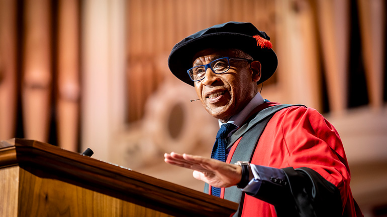 Lord Simon Woolley in academic robes speaking at a lectern during a graduation ceremony.