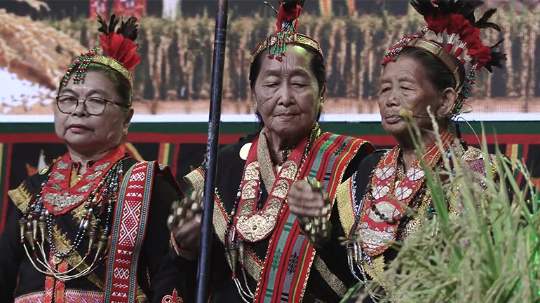 Three Lotud priestesses wearing traditional ceremonial dress and ornate feathered headdresses during a cultural performance.