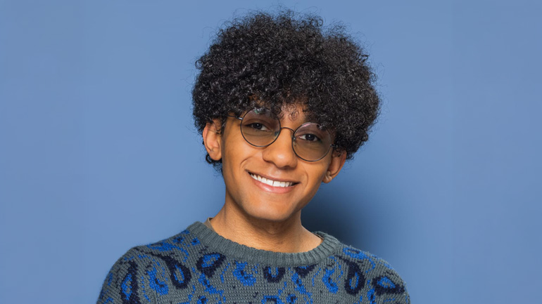 Portrait of a Shakeer Sayani wearing glasses and a patterned jumper against a blue background.