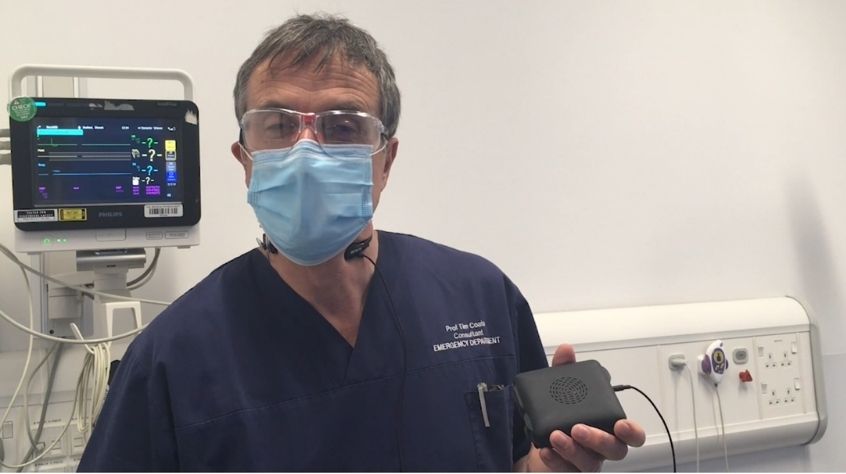 A doctor wearing a face mask and protective glasses holds up a small electrical device.