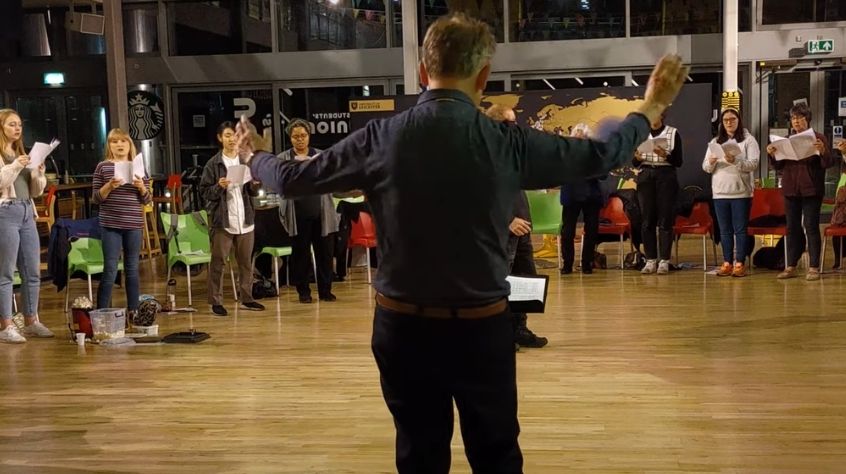 An individual, with back to the camera, conducts a choir of students.