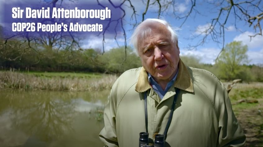 Sir David Attenborough in front of a large pond and blue sky.