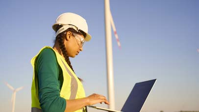Female engineer wearing hard hat and reflective green jacket standing with laptop against wind turbine and setting it up.