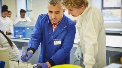 Lecturer and student working on an experiment in a lab with other students seen in the background.