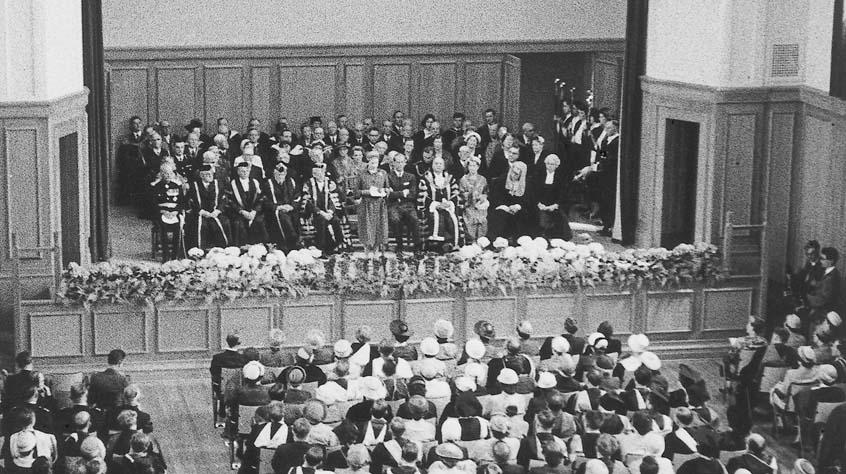 Her Majesty The Queen speaking on stage, surrounded by senior academics