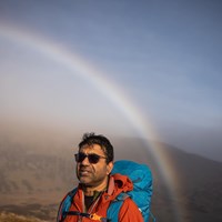 "Mohammed Dhalech is in the mountains wearing an orange jacket and blue rucksack and a rainbow over him. wearing dark glasses.,"