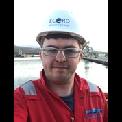 A headshot picture of Andrew McIntyre in red overalls, white hard hat, safety glasses aboard a ship  