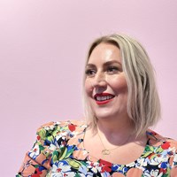 A white woman with a blonde bob, wearing red lipstick and a colourful flowery top looks out to the left, against a pink wall