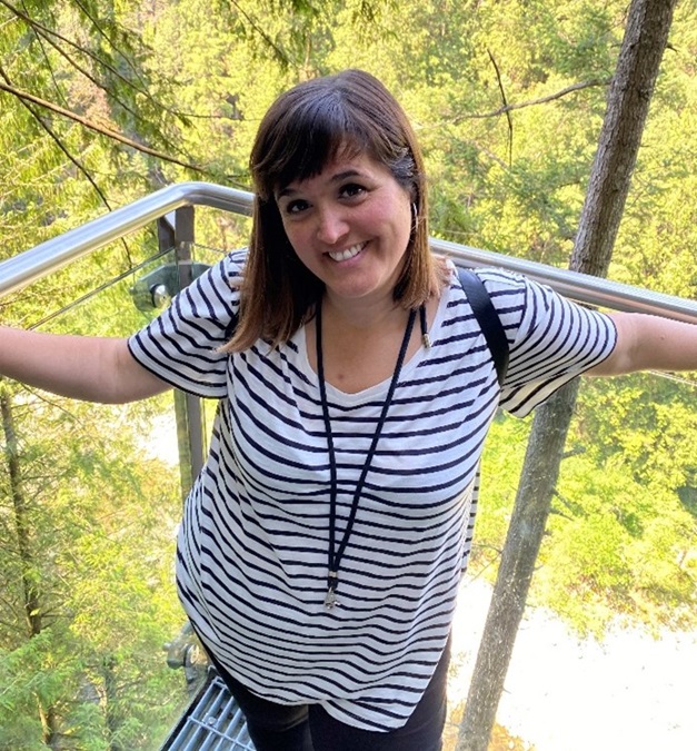 Dr Patricia Rodríguez-Maciá, wearing a horizontal-striped shirt, standing on a balcony in front of a green background. Patricia is smiling.