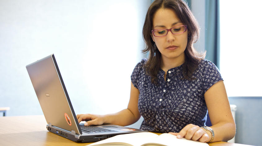 Student in front of open laptop looking at a book.