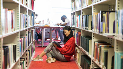 Student sitting on the floor of the library reading a book.