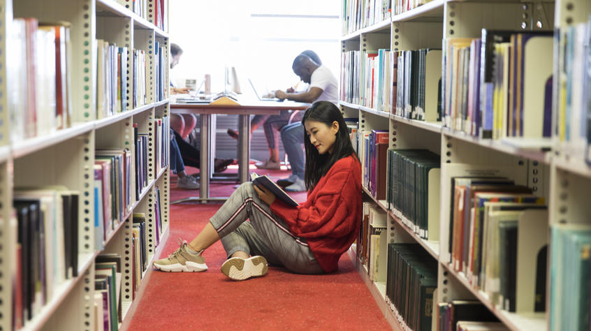 Student sitting on the floor of the library reading a book.