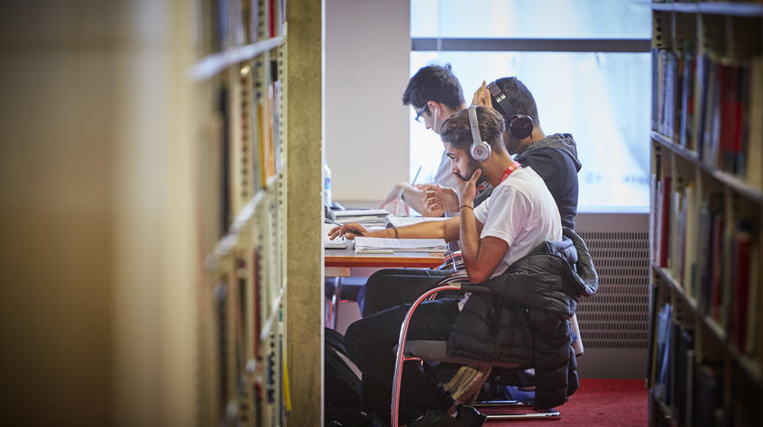 Students working at a table in the library at the end of bookshelves.