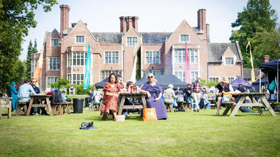 People sit at picnic tables on a lawn in front of a large brick building during a sunny outdoor event.