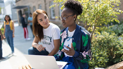 Two students sitting and talking outside the David Wilson Library, with a laptop on one of their knees.
