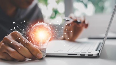 Person sat working on a laptop, with a lit up lightbulb in their right hand.