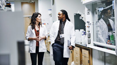 two people wearing lab coats walking through a lab and talking