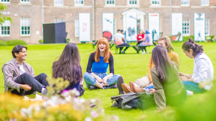 Students sitting on the grass, talking and laughing outside the Fielding Johnson Building on Leicester campus.