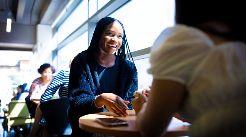 Student sitting in the library cafe, talking and laughing with a friend.