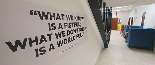 Photograph of a sign with the quote "What we know is a fistful; what we don't know is a world full" a Sri Lankan proverb, is printed on the wall. There are some soft furnishing booths in the background.